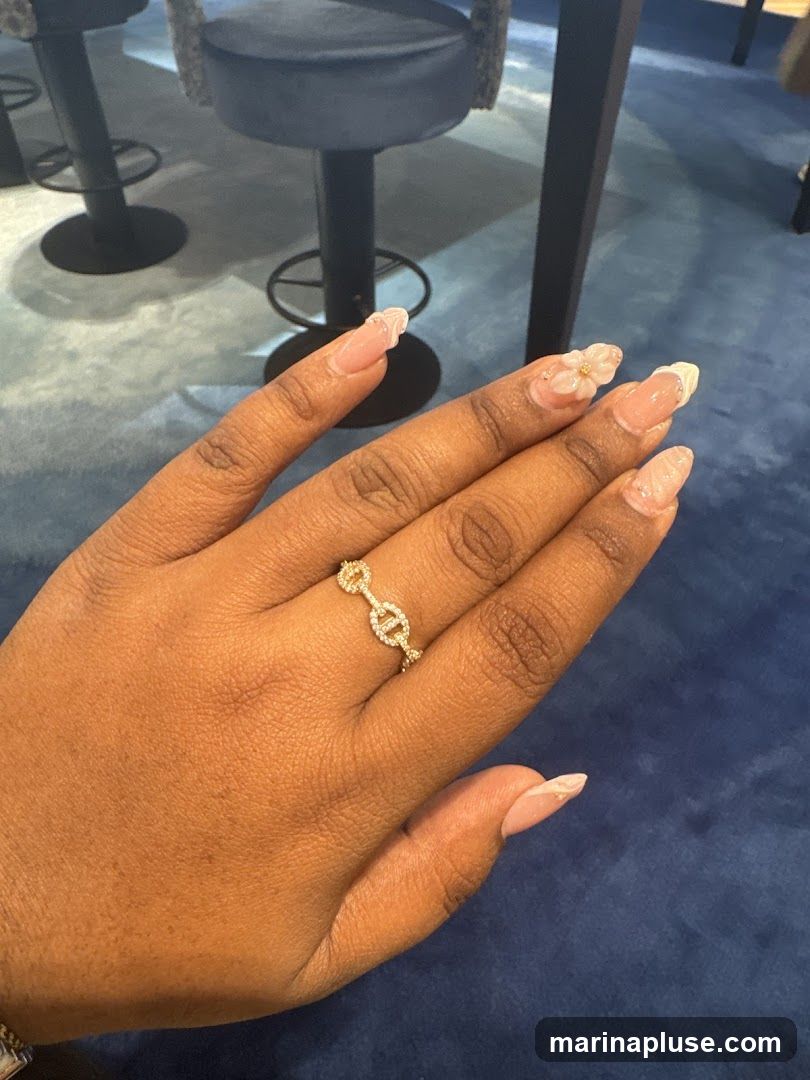 A close-up shot of a person's hand adorned with a gold ring featuring interlocking links and small diamonds. The fingernails are decorated with a French manicure style, featuring white tips and subtle floral or abstract designs, with one nail featuring a small 3D flower embellishment. The background appears to be a blue upholstered surface, possibly a chair or sofa, with dark legs of stools visible.