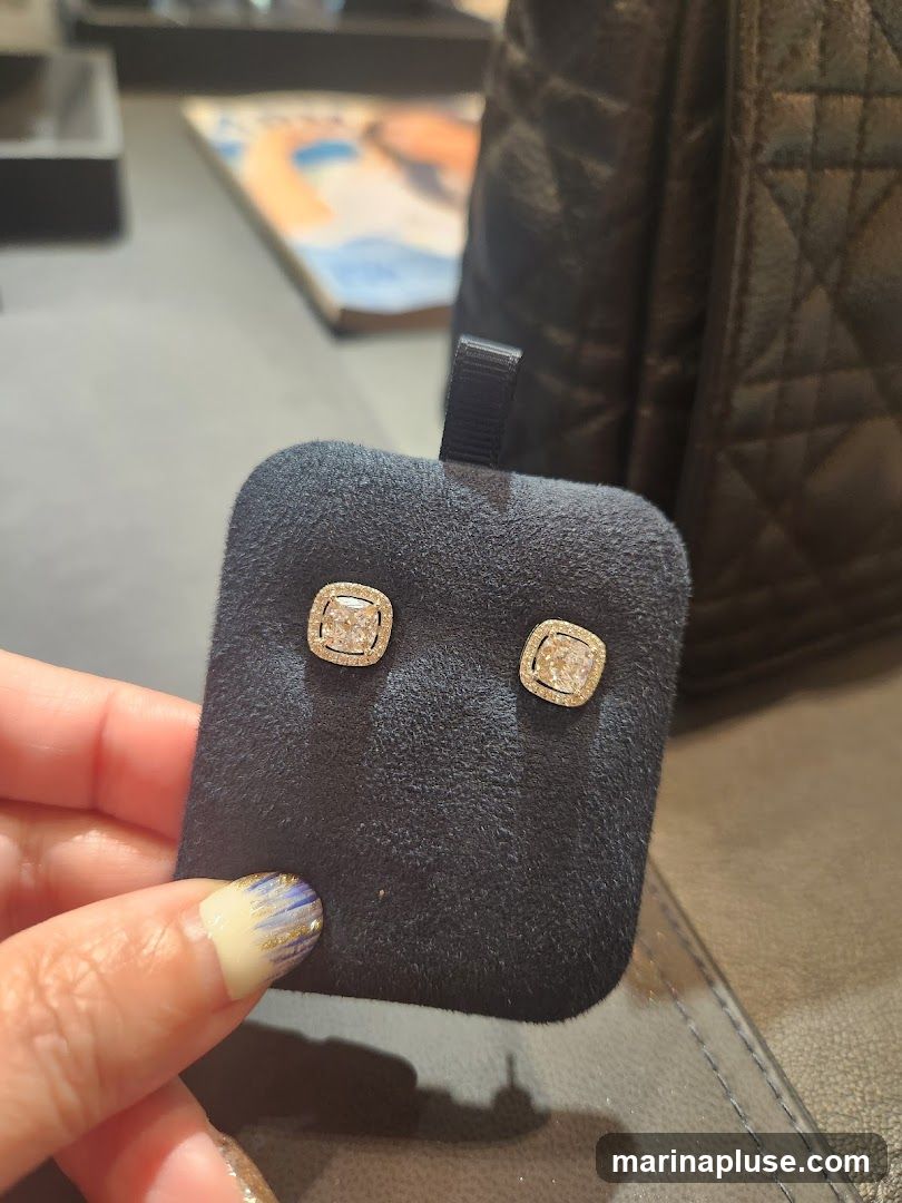A hand with decorated nails holds a black display stand with two cushion-cut diamond stud earrings. The earrings are set in gold and surrounded by smaller diamonds. In the background, a quilted black bag and a magazine are visible.