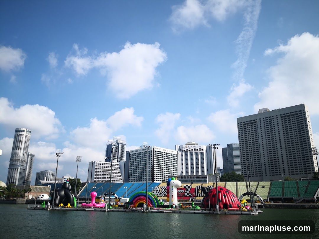 A collection of large inflatable attractions, including a panda, a unicorn, a ladybug, and a rainbow arch, are set up on a floating platform on the water. In the background, a modern city skyline with tall buildings is visible under a bright blue sky with scattered clouds. Stadium seating can be seen behind the inflatables.