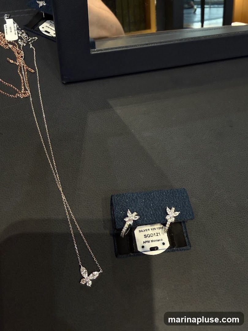 A close-up shot of a delicate silver necklace with a butterfly pendant adorned with clear stones. Next to it, a small blue pouch holds a pair of matching butterfly stud earrings, also embellished with stones. A price tag is attached to the earring pouch, reading "SILVER 925-1000 SGD121 APM Monaco". The items are displayed on a dark, textured surface, possibly a jewelry display. A reflection of a person and a bookshelf is visible in a dark frame in the background.