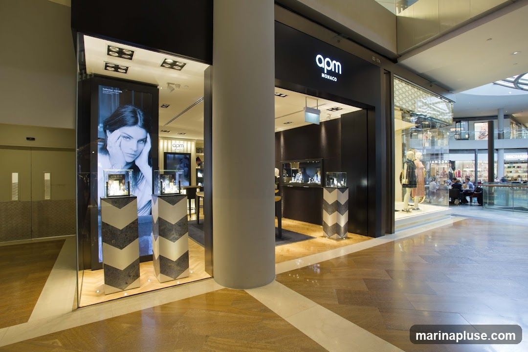 The storefront of Apm Monaco, a jewelry store, is visible in a mall. The entrance features a dark facade with the store's logo illuminated in white. To the left, a large display window showcases a model's portrait and jewelry on stands. Inside, display cases with jewelry are visible, and mannequins are dressed in the window to the right. Other stores and shoppers are visible in the background.