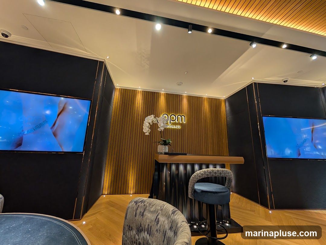 A modern reception area with a wooden paneled wall featuring a logo that reads 'apm MONACO'. Two large screens display abstract blue imagery. A reception desk with a wooden counter is present, along with a blue velvet bar stool. The floor is made of herringbone wood.