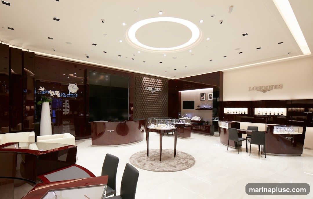 The interior of a luxurious Longines watch store. The space features polished floors, dark wood display cases, and elegant seating areas. A large screen is visible behind a reception desk, and the Longines logo is prominently displayed on a textured wall and above a display counter. Various watches are showcased in glass display cases throughout the store.