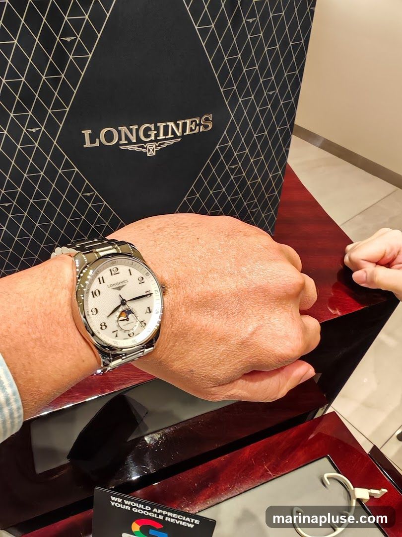 A person is wearing a Longines watch with a white dial and a moon phase complication. The watch is displayed on a wooden surface, with a Longines branded bag in the background. A card requesting a Google review is also visible.