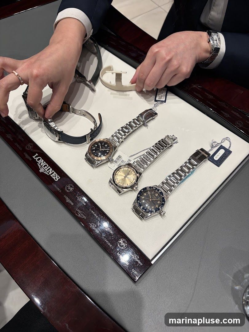 A close-up view of several Longines watches laid out on a display tray. Two individuals' hands are visible, one adjusting a watch and the other holding a watch strap. The watches have metal bracelets and various dial colors, including black and gold. Tags are attached to some of the watches. The display tray has the Longines logo repeated along its edge.