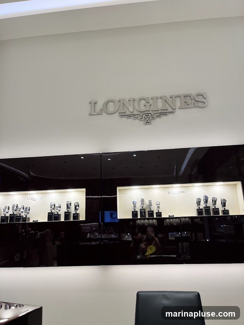 A display case featuring Longines watches. The Longines logo is prominently displayed on the wall above the display. Several watches are showcased on stands within illuminated compartments. Reflections of people and the interior of the store are visible on the glossy black surface of the display case.