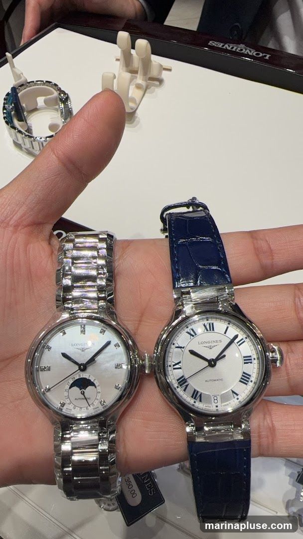 A hand holds two Longines watches. The watch on the left has a silver band and a white dial with a moon phase complication. The watch on the right has a blue leather strap and a white dial with Roman numerals. A price tag of 350,00 is visible on the left watch. In the background, a display case with the Longines logo is visible.