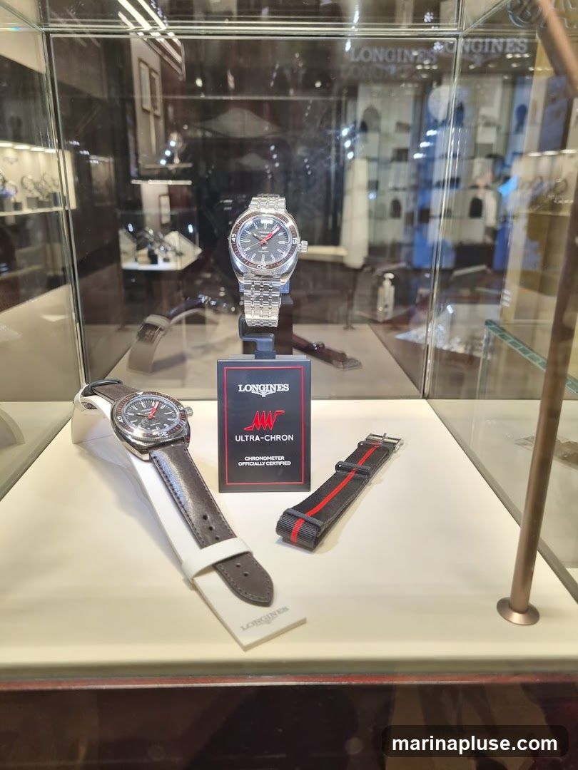 A display case featuring two Longines watches. One watch is on a metal bracelet, and the other is on a dark leather strap. A red and black fabric watch strap is also displayed. A sign in the center reads "LONGINES ULTRA-CHRON CHRONOMETER OFFICIALLY CERTIFIED". The background shows a blurred interior of a store with more watches and displays.