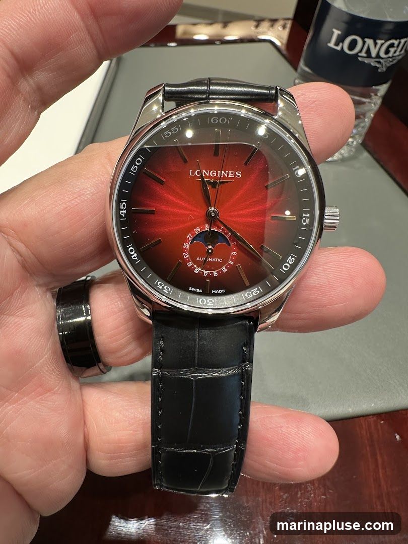 A close-up shot of a Longines watch with a red sunburst dial and a black leather strap. The watch features a moon phase complication at the 6 o'clock position. A bottle with the Longines logo is visible in the background.