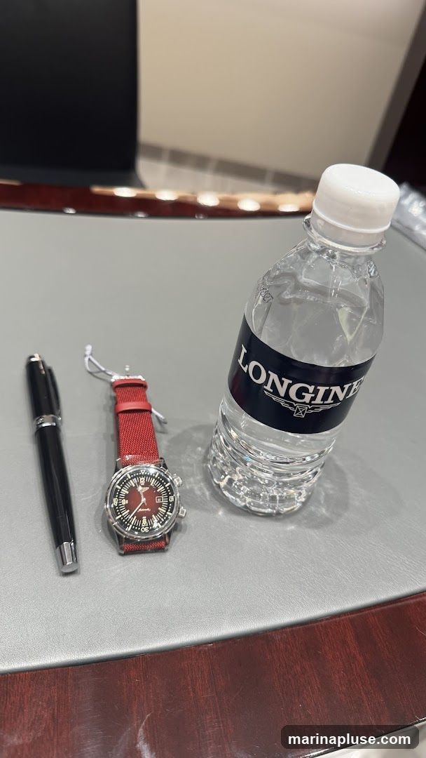 A black pen, a red-strapped Longines watch, and a bottle of water with a Longines label are placed on a grey surface with a wooden trim.