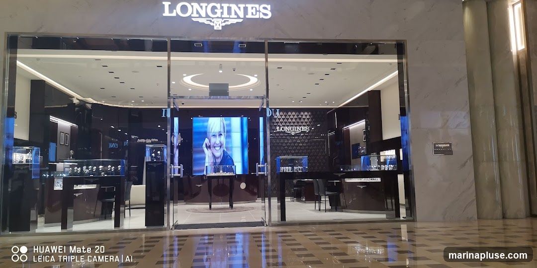 The exterior of a Longines watch store. The store has a large glass facade with the Longines logo and name prominently displayed above the entrance. Inside, display cases showcase watches, and a large screen displays an advertisement featuring a smiling woman. The interior is modern and sleek, with dark wood and polished surfaces.