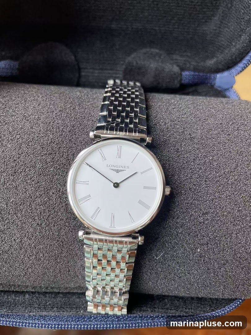 A close-up shot of a Longines watch with a white face and Roman numerals. The watch has a silver metal band and is resting on a textured gray surface inside a blue case.