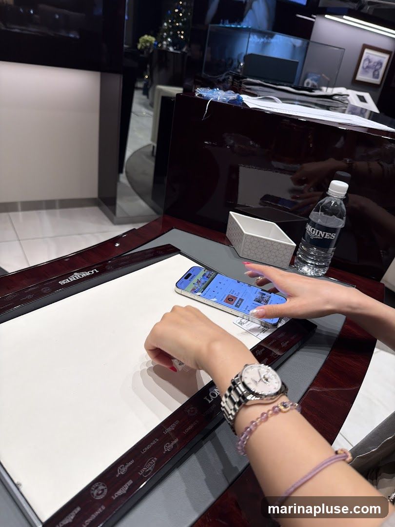 A person's hands are visible, one holding a smartphone displaying a website with images and text, possibly a catalog or online store. The other hand is resting on a white surface. A watch is worn on the wrist. The background shows a display case with a Christmas tree and other items, suggesting a retail environment. A bottle of water is also on the table.