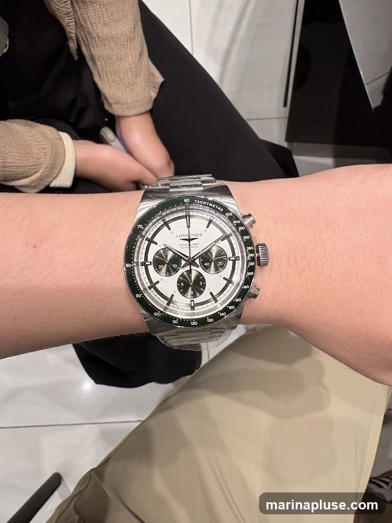A close-up shot of a person wearing a Longines Conquest watch on their wrist. The watch has a silver dial with three sub-dials and a green tachymeter bezel. The watch band is made of stainless steel. The background shows parts of people sitting down.