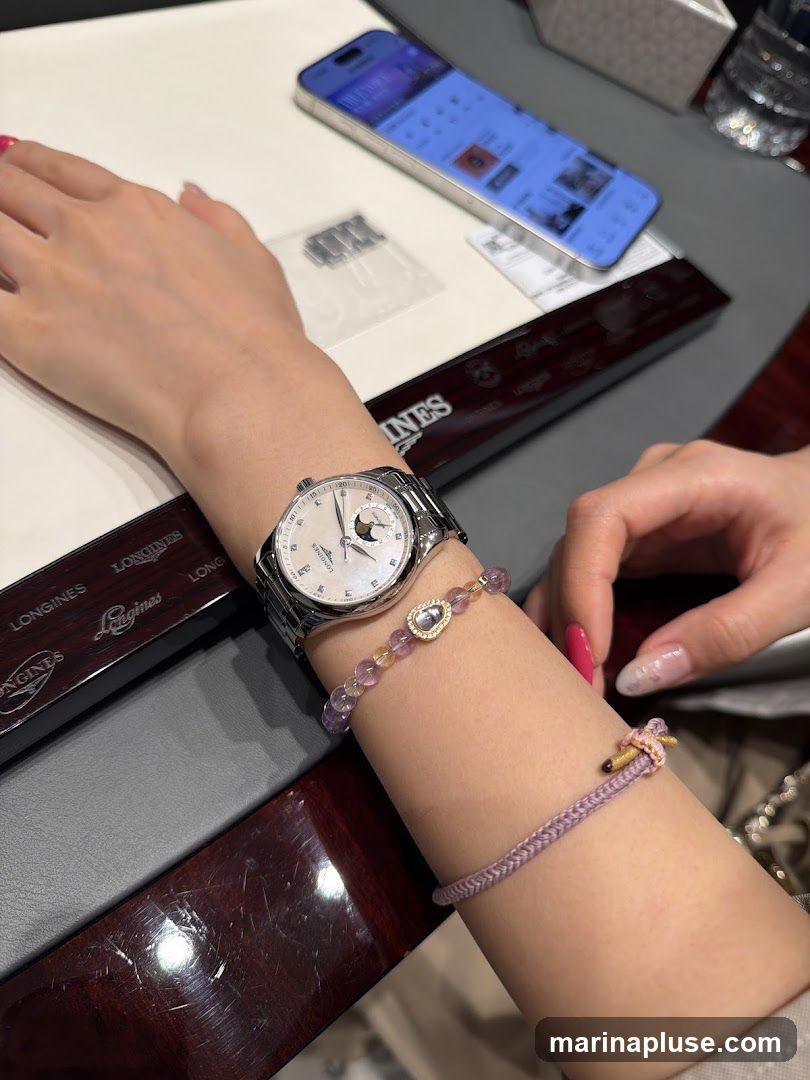 A close-up shot of a woman's arm wearing a silver watch with a white face and a moon phase complication. She also has two bracelets on her wrist: one with purple and yellow beads and a central pendant, and another braided purple string bracelet. A smartphone is visible in the background, displaying a screen with various icons and images. A box with the Longines logo is also present.