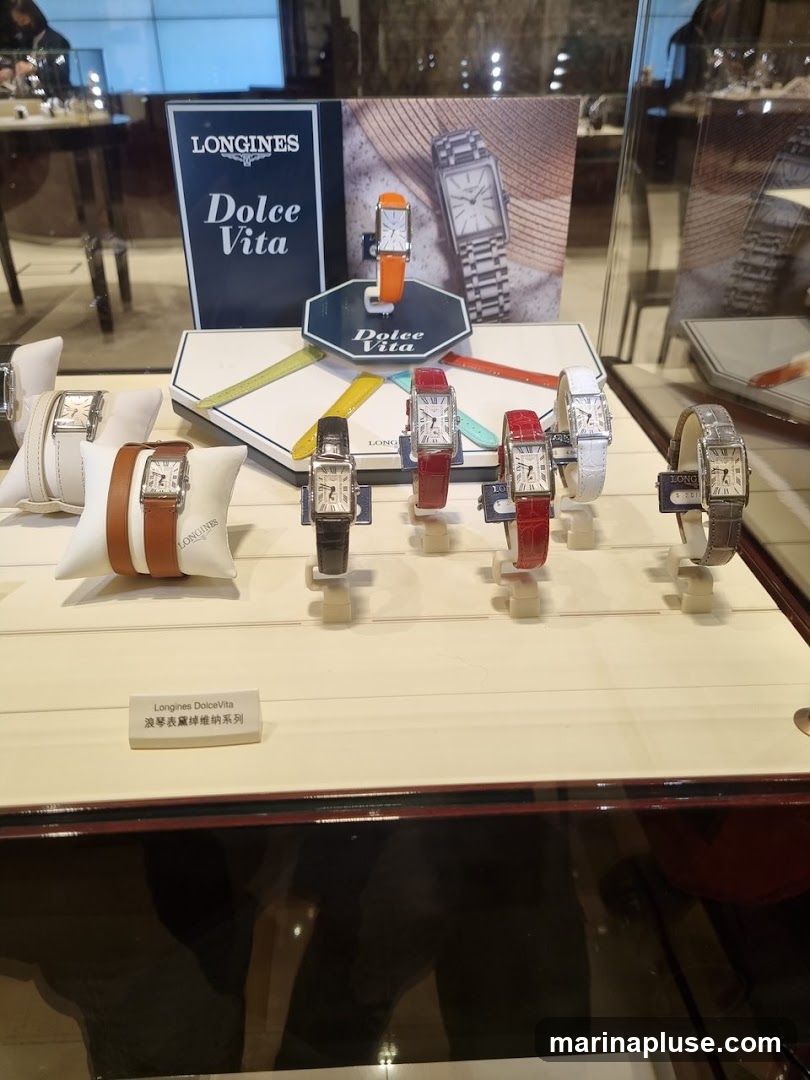 A display case featuring several Longines Dolce Vita watches. The watches have rectangular faces and come with various colored straps, including brown leather, black leather, red, white, and grey. A promotional sign for the Longines Dolce Vita collection is visible in the background.