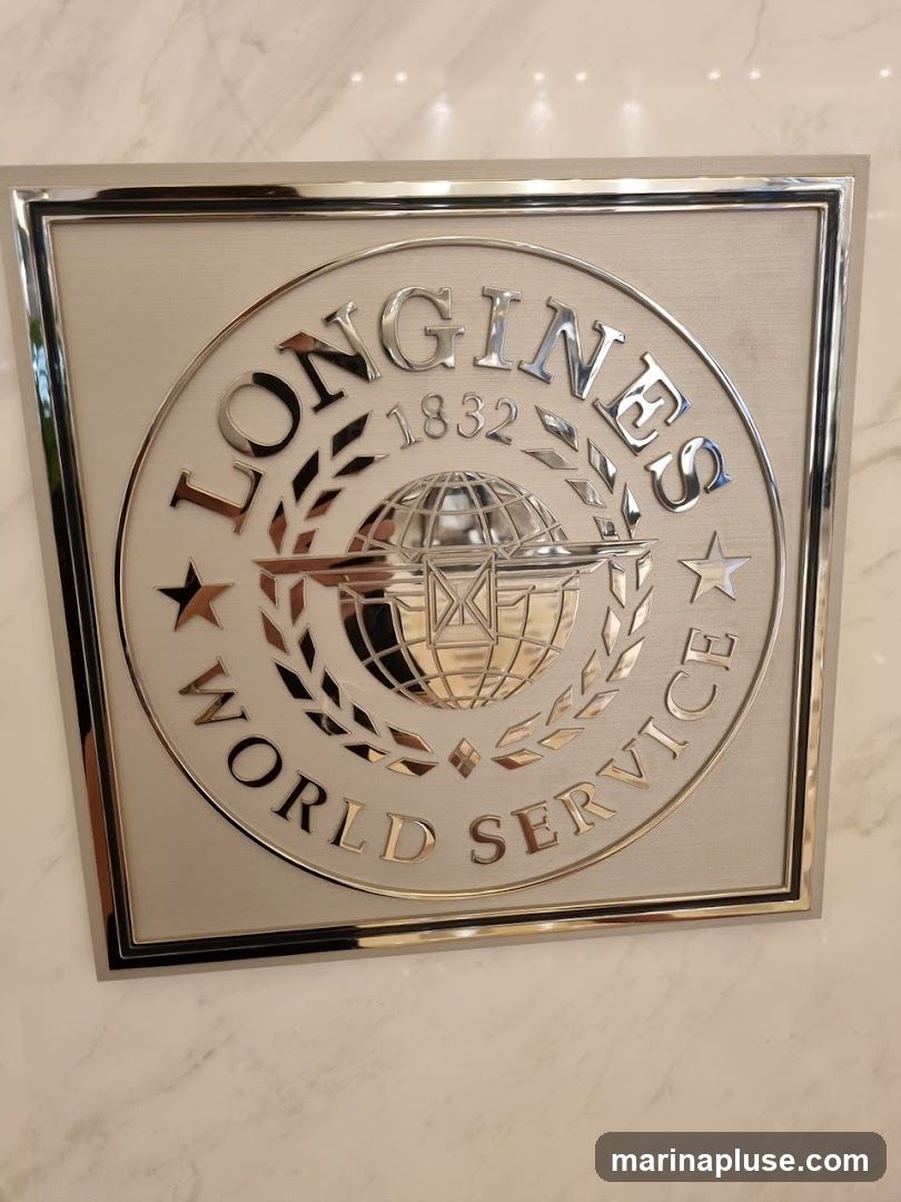 A close-up, angled shot of a metallic emblem for Longines World Service. The emblem is circular with the brand name "LONGINES" arched above and "WORLD SERVICE" arched below. In the center is a globe with wings, and the year "1832" is visible. The emblem is set within a square frame with a polished silver border. The background appears to be a marble-like surface.