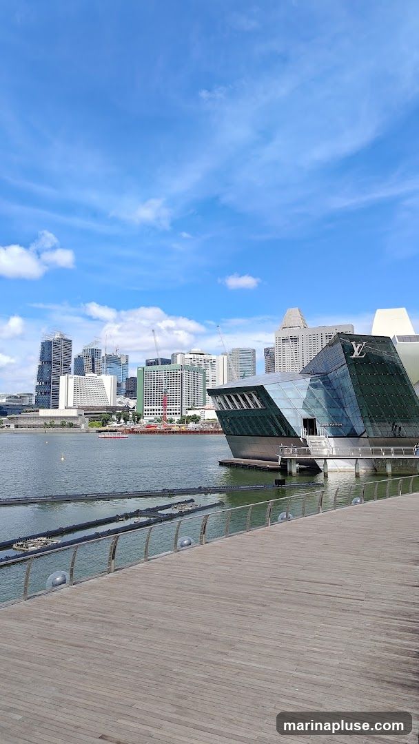 A modern, angular glass building with the Louis Vuitton logo prominently displayed on its facade, situated on the water's edge. The building has a sleek, contemporary design with reflective glass panels. In the background, a cityscape with various tall buildings is visible under a bright blue sky with scattered clouds. A wooden boardwalk runs along the foreground, leading towards the building.