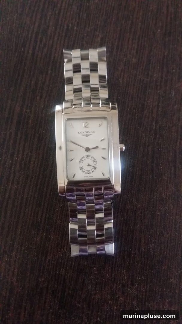 A rectangular silver Longines watch with a white face and a silver band is shown against a dark, textured background. The watch face has Roman numerals for the 12 o'clock position and small markers for other hours. A subdial is visible at the 6 o'clock position. The brand name "LONGINES" is printed below the 12 o'clock marker.