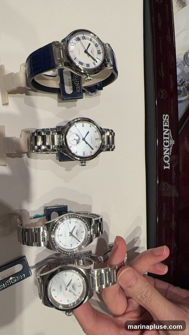 A display of four Longines watches. The top watch has a dark blue leather strap and a white dial with Roman numerals. The second watch has a silver metal bracelet and a white dial with a sub-dial. The third watch has a silver metal bracelet and a white dial with diamond hour markers. The bottom watch, being held by a hand, has a silver metal bracelet and a white dial. A dark box with repeated "LONGINES" branding is visible on the right. Price tags are visible on some watches.