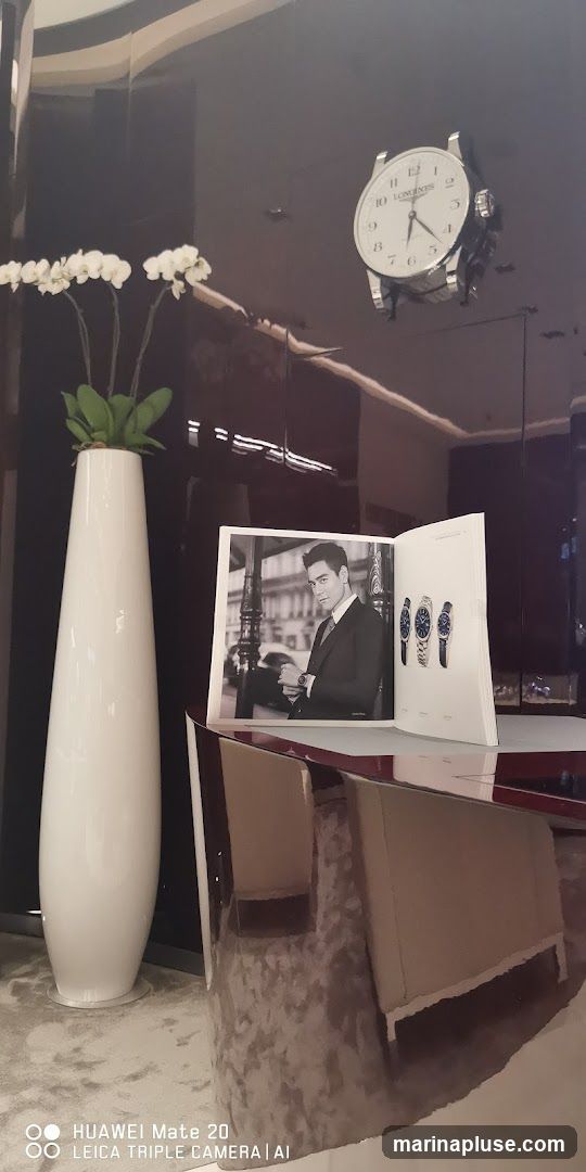 A display featuring a large clock with the Longines logo, a tall white vase with white orchids, and an open brochure showcasing a man in a suit and several watches. The scene appears to be inside a store or showroom.