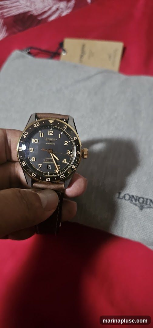 A person is holding a Longines automatic chronometer watch with a dark dial, gold bezel, and brown leather strap. The watch has a GMT function indicated by the 24-hour scale on the bezel and a separate hand. In the background, a grey fabric with the Longines logo is visible, along with a brown tag. The overall impression is of a luxury timepiece.