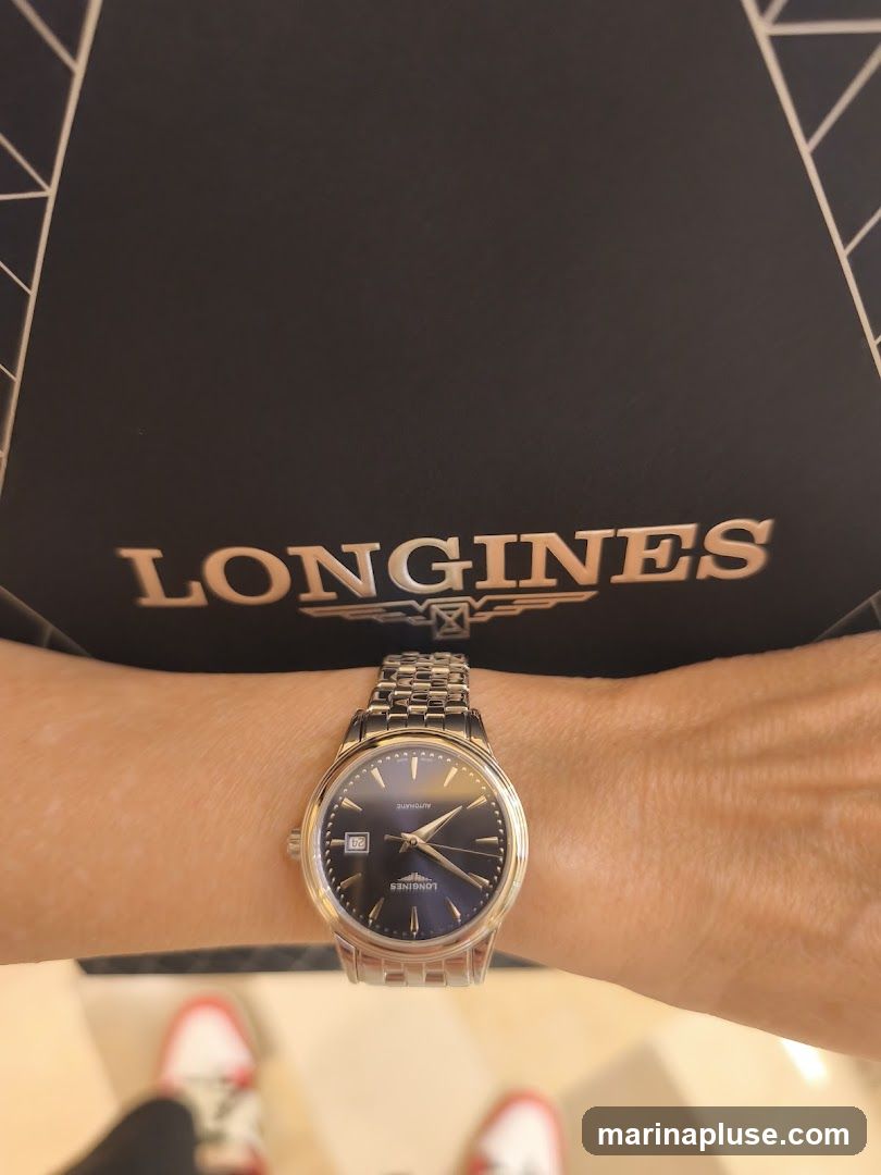 A person's wrist is adorned with a silver Longines watch with a dark blue dial. The watch features a date window at the 3 o'clock position and a metal link bracelet. Behind the wrist, a black surface with the rose gold logo "LONGINES" is visible. The bottom of the image shows a blurred view of red and white sneakers.
