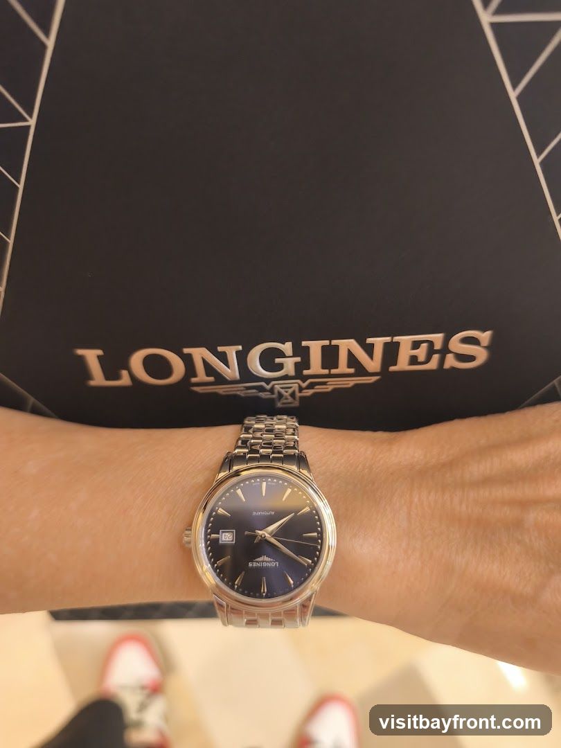 A person's wrist is adorned with a silver Longines watch with a dark blue dial. The watch features a date window at the 3 o'clock position and a metal link bracelet. Behind the wrist, a black surface with the rose gold logo "LONGINES" is visible. The bottom of the image shows a blurred view of red and white sneakers.