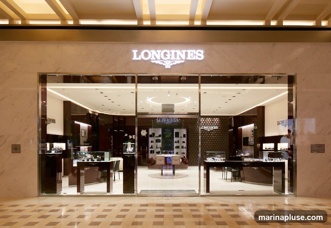 A well-lit, modern storefront for a luxury watch brand. The facade features large glass windows displaying watches in illuminated cases. Above the entrance, the brand name "LONGINES" is prominently displayed in illuminated white lettering with its logo. The interior is visible through the glass, showing sleek display cabinets and a polished floor.