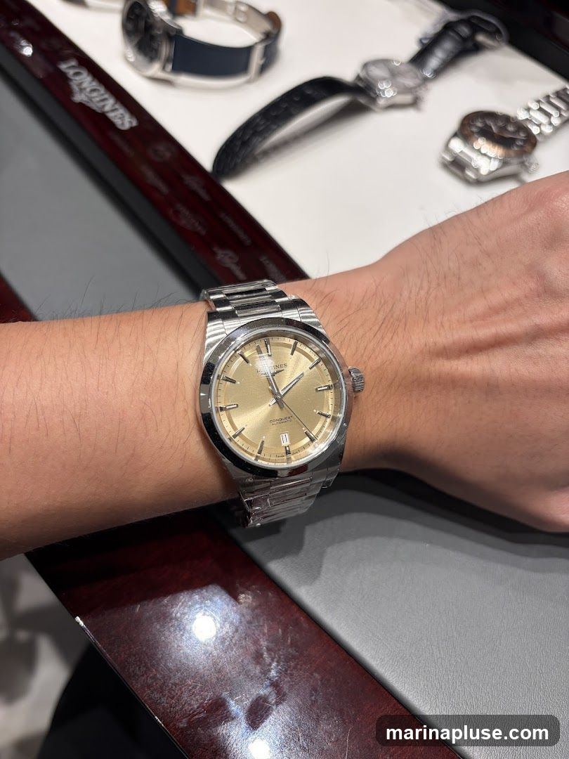 A close-up shot of a person's wrist wearing a silver Longines watch with a champagne-colored dial. The watch has a metal bracelet and appears to be a Conquest Automatic model. In the background, other watches are displayed on a dark wooden surface with the Longines logo visible.