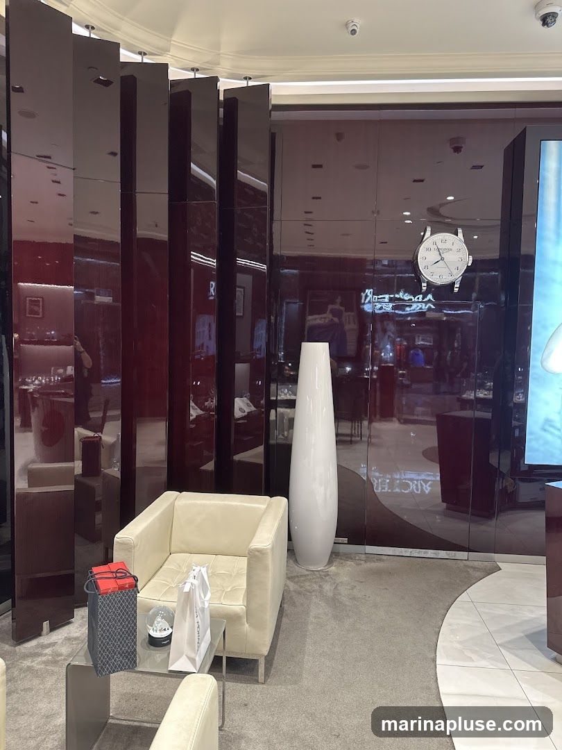 A plush cream-colored armchair sits in a well-lit, modern interior, possibly a luxury watch store. A glass-topped table next to the armchair holds two shopping bags and a snow globe. The background features a wall with reflective panels, displaying a clock and what appears to be a digital advertisement. Tall, slender white vases add to the sophisticated ambiance.