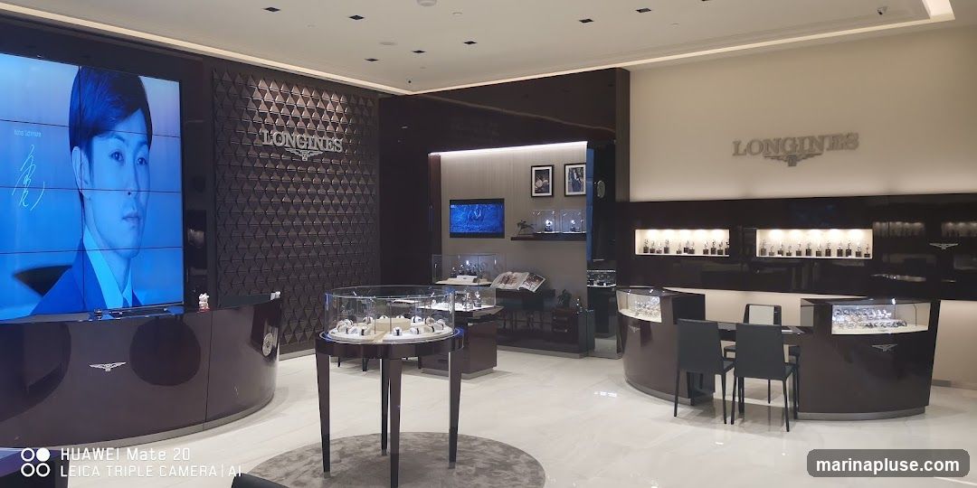 The interior of a Longines watch store, featuring display cases filled with watches, a large screen displaying a portrait of a man, and prominent Longines branding on the walls. The store has a modern and luxurious design with polished floors and dark wood accents.