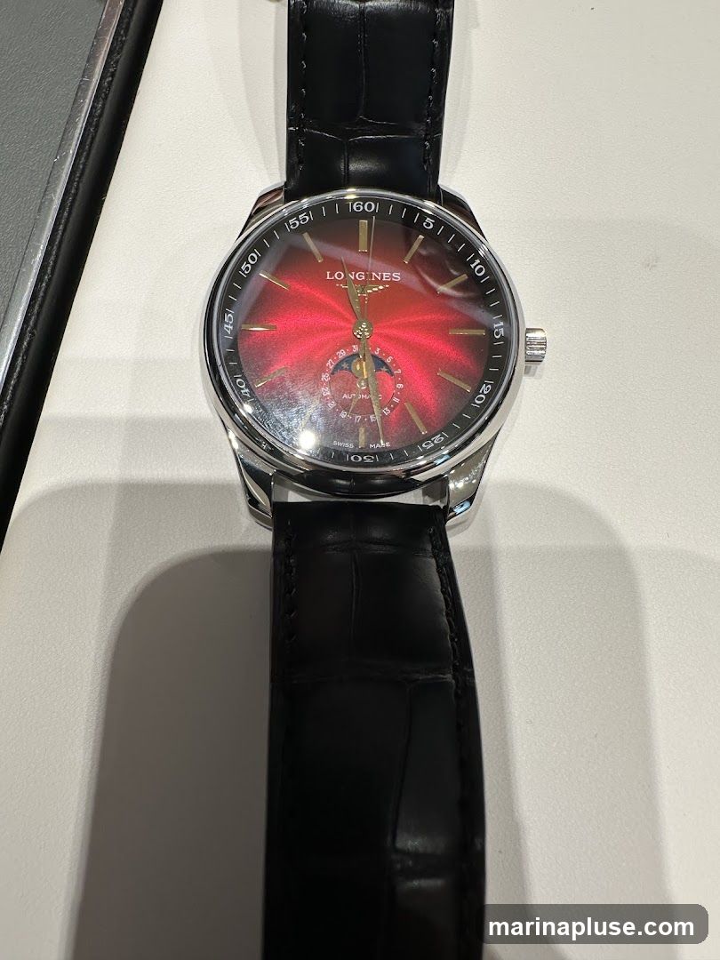 A close-up shot of a Longines watch with a red sunburst dial and a black leather strap. The watch face features gold-toned hour markers, a date subdial, and the Longines logo. The watch is displayed on a white surface.