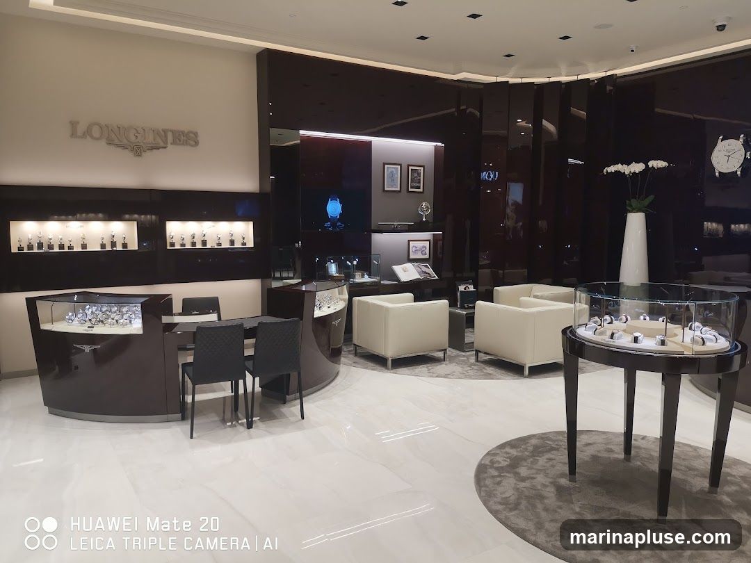 The interior of a Longines watch store. The store features a large display of watches on shelves and in glass cases. There are also seating areas with plush armchairs. The store is well-lit and has a modern, luxurious design.