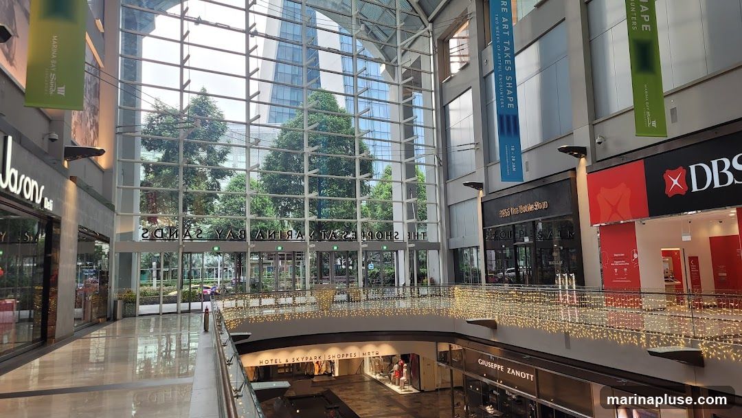 An interior view of the Marina Bay Sands Shoppes, featuring a large glass atrium, storefronts, and decorative lights. Visible storefronts include Jason's Deli, The Bottle Shop, DBS bank, and Giuseppe Zanotti. Banners advertising "Art Takes Shape" and "Marina Bay Sands" are displayed.