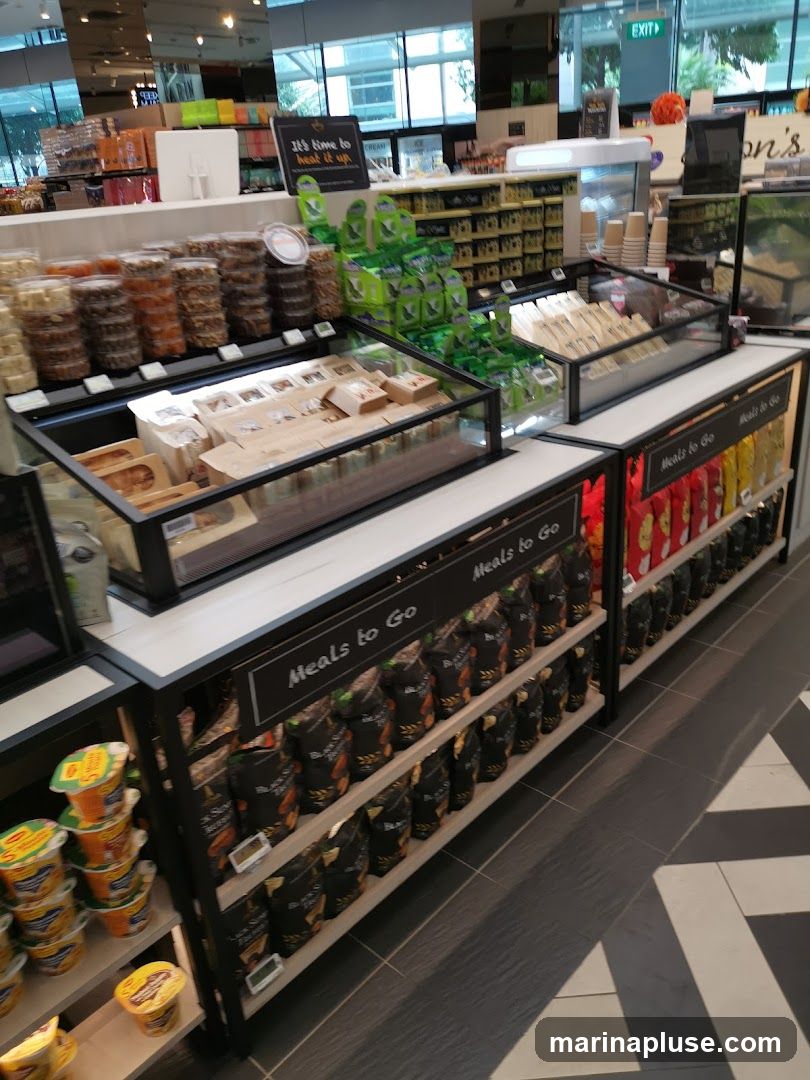A display of pre-packaged meals and snacks in a store. The shelves are stocked with various items, including bags of chips, instant noodle cups, and items in clear containers. A sign reads "Meals to Go" above a selection of bagged snacks. Another sign says "It's time to heat it up.". In the background, more shelves of products are visible, along with an exit sign.