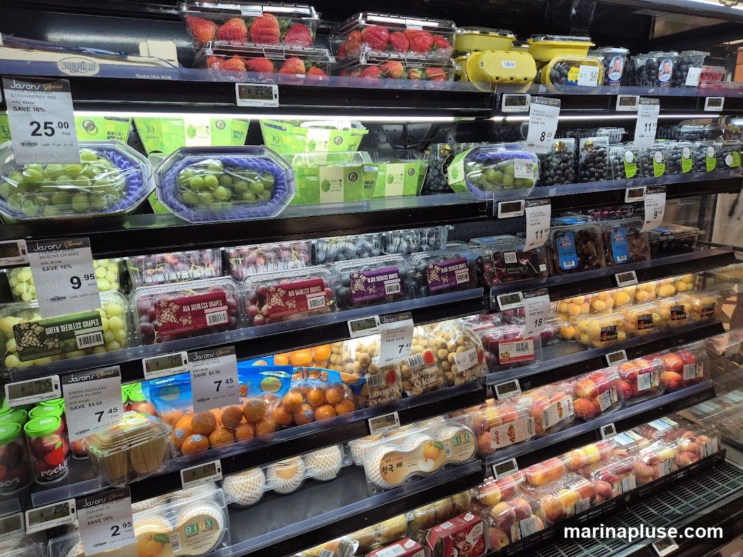 A refrigerated display case filled with various fruits, including strawberries, green and red seedless grapes, blueberries, oranges, peaches, and longans. Many of the fruits are packaged in clear plastic containers with price and discount labels.
