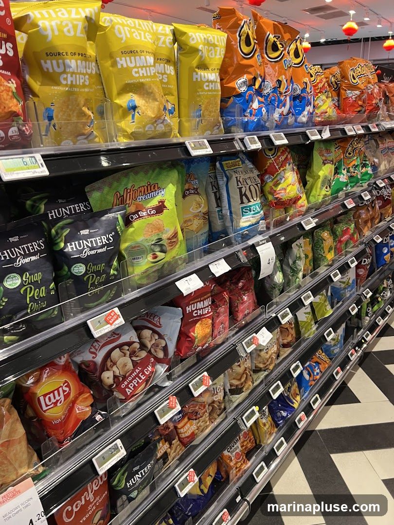 A brightly lit aisle in a store displays a wide variety of snack bags on multiple shelves. The snacks include various brands and flavors of chips, such as Hummus Chips, Lay's, and Hunter's Gourmet. Some bags are large and colorful, while others are smaller and more subdued. Price tags are visible on some of the shelves. The flooring has a black and white striped pattern.