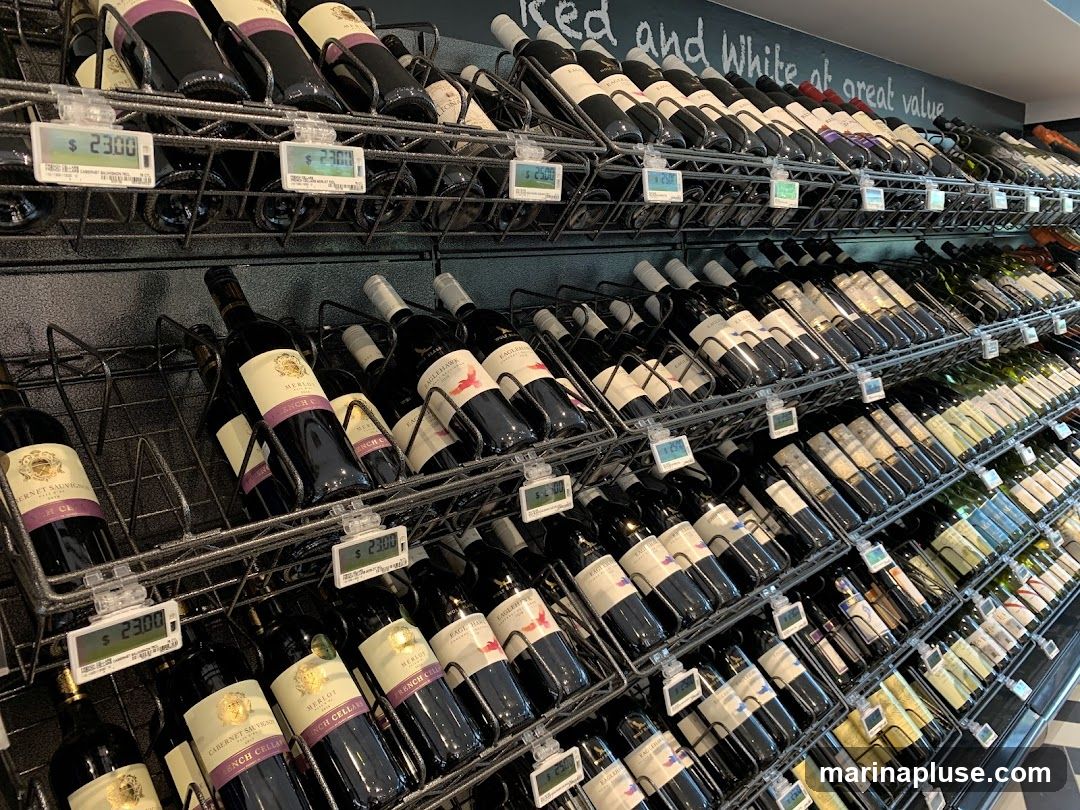 A wide selection of wine bottles are displayed on tiered shelves in a store. Digital price tags are visible in front of some bottles. The shelves are organized with bottles facing forward, and some bottles are tilted. A chalkboard sign above the top shelf reads "Red and White at great value".