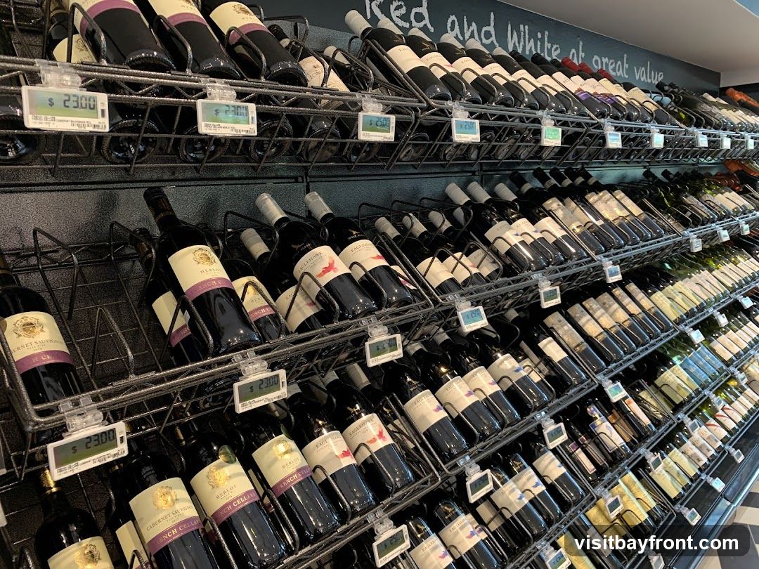 A wide selection of wine bottles are displayed on tiered shelves in a store. Digital price tags are visible in front of some bottles. The shelves are organized with bottles facing forward, and some bottles are tilted. A chalkboard sign above the top shelf reads "Red and White at great value".