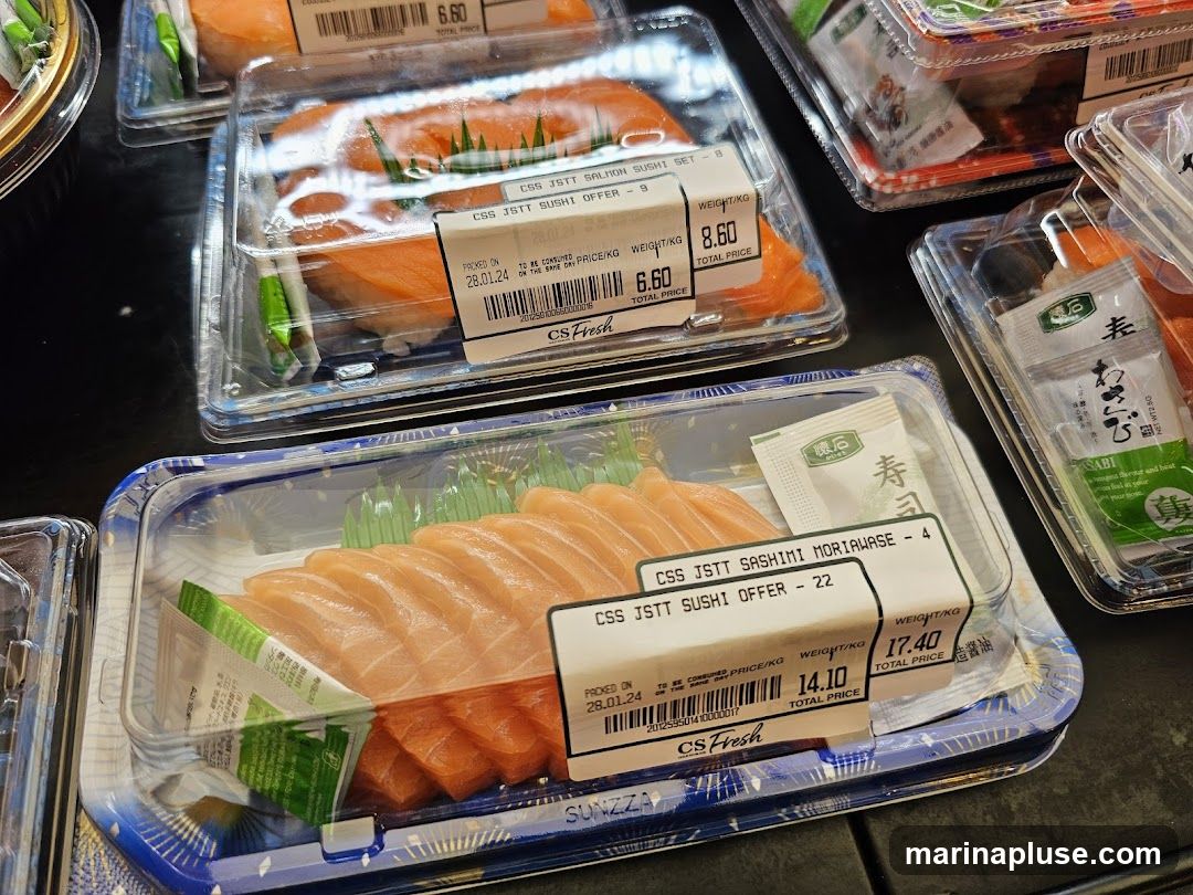 A close-up shot of pre-packaged sushi and sashimi sets displayed in clear plastic containers. The salmon sushi sets and sashimi moriawase are neatly arranged, with small packets of wasabi and soy sauce included. Labels indicate product names, prices, and packing dates. The CS Fresh logo is visible on some labels.