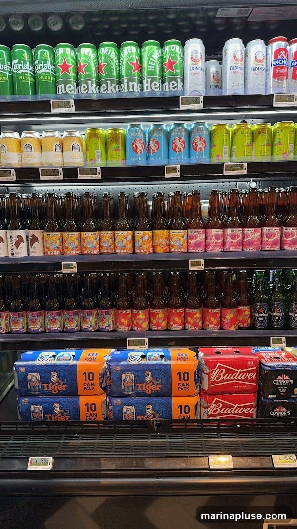 A refrigerated display case filled with various brands of beer in cans and bottles. The top shelf shows cans of Carlsberg, Heineken, and Erdinger. The second shelf has cans of what appear to be craft beers and lemonade. The third and fourth shelves are stocked with bottles of various beers, some with colorful labels. The bottom shelf displays multi-packs of Tiger, Budweiser, and Connor's Stout Porter. Price tags are visible below some of the products.