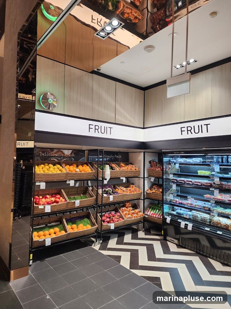 A well-lit section of a store dedicated to fruits, with wooden crates filled with various fruits like oranges, lemons, and apples. A refrigerated display case filled with packaged goods is visible on the right. The word "FRUIT" is prominently displayed on the wall above the fruit displays. The floor has a black and white chevron pattern.
