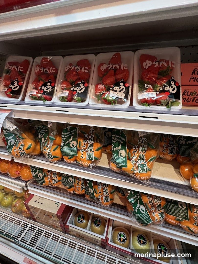 A refrigerated display case filled with various fruits. The top shelf shows several packages of strawberries, each with a black bear character and Japanese text. The middle shelves contain bags of oranges labeled "Tamura Mikan" and "Komina Farm." The bottom shelf displays boxes of what appear to be kiwis, some with "We Fresh" stickers.