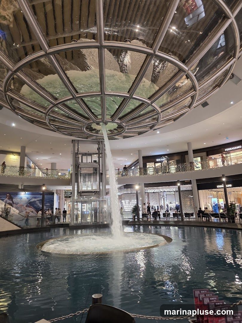 An indoor shopping mall features a large, multi-story water feature that cascades from a circular structure at the ceiling into a pool below. Surrounding the pool are various retail stores with illuminated storefronts, including Emporio Armani and Stone Island. People are seen browsing the shops and sitting at tables near the water. The mall has multiple levels with balconies and decorative lighting.
