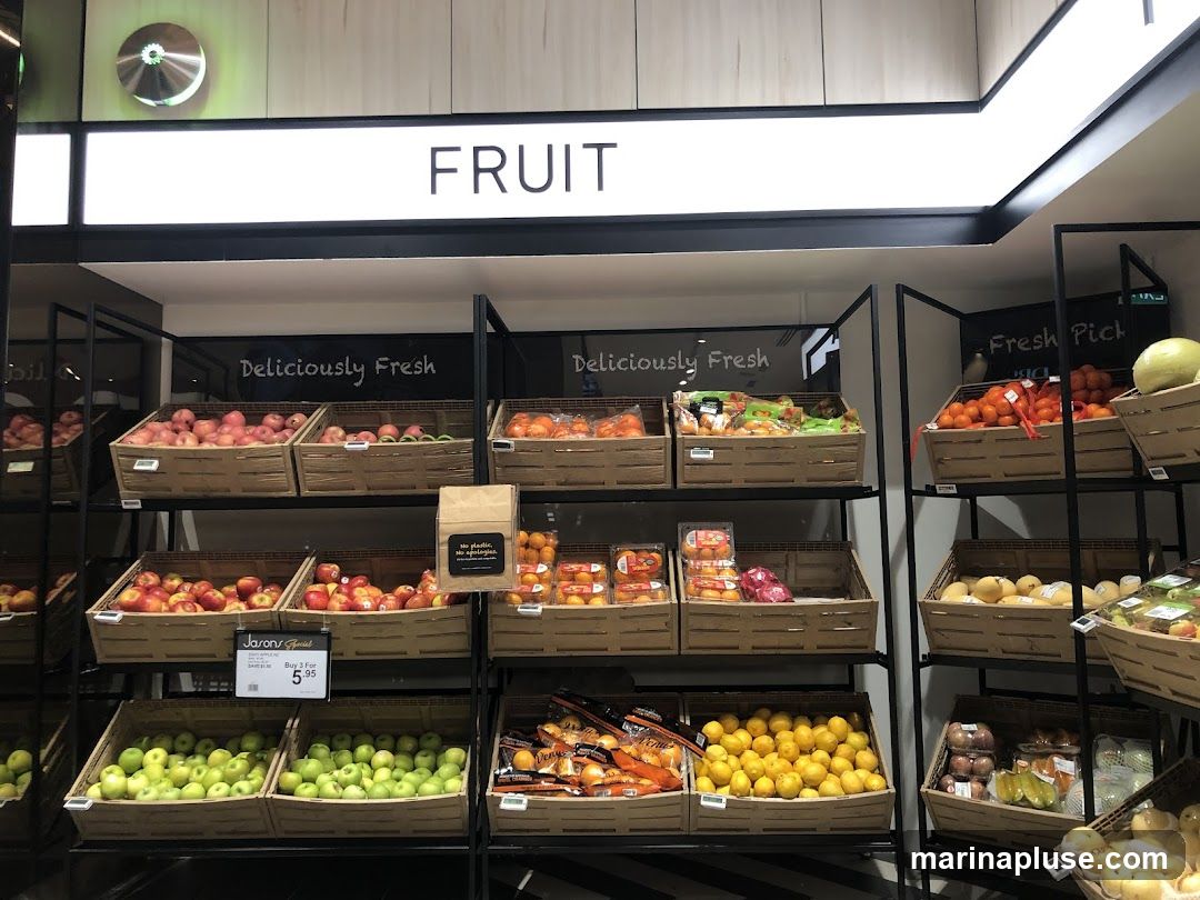 A grocery store display featuring various fruits in baskets. The sign above reads "FRUIT". Below, "Deliciously Fresh" is written. Fruits visible include apples, oranges, lemons, and other produce. A sign indicates "Jason's Special" with a price of "Buy 3 For $5.95". Another sign says "No plastic, no apologies."