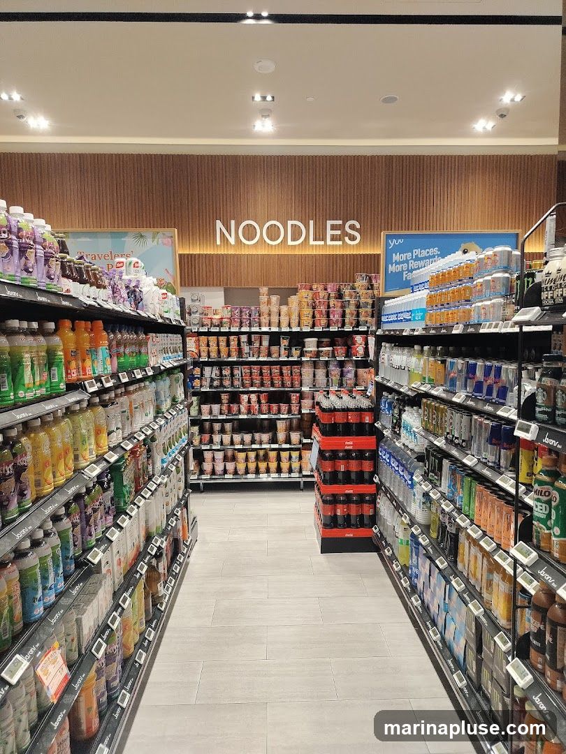 An aisle in a store with shelves stocked with various beverages and instant noodles. The word "NOODLES" is prominently displayed on the wall above a section of instant noodle cups. On the left, shelves are filled with bottled drinks, and on the right, more bottled drinks and cans are visible.
