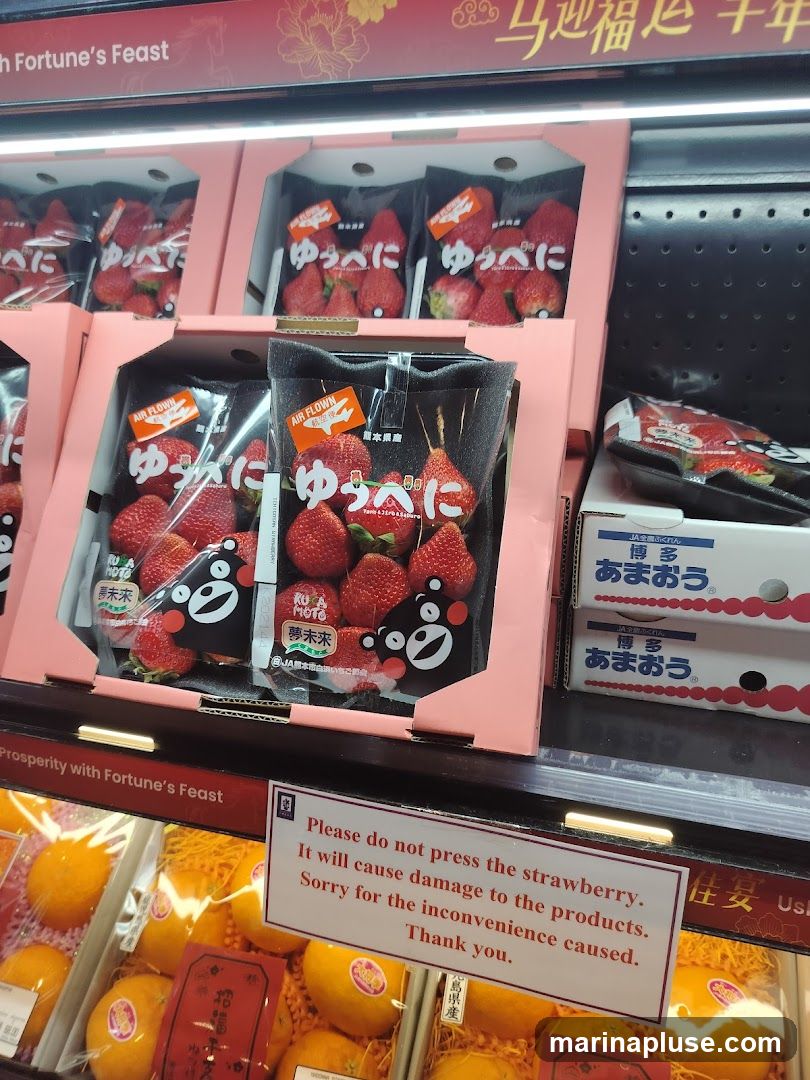 A display of packaged strawberries and oranges in a store. The strawberries are in pink boxes and plastic packaging, some with cartoon characters. A sign reads "Please do not press the strawberry. It will cause damage to the products. Sorry for the inconvenience caused. Thank you." Below the strawberries, oranges are displayed in clear plastic packaging. Decorative text in Chinese and English is visible at the top and on a sign above the oranges.