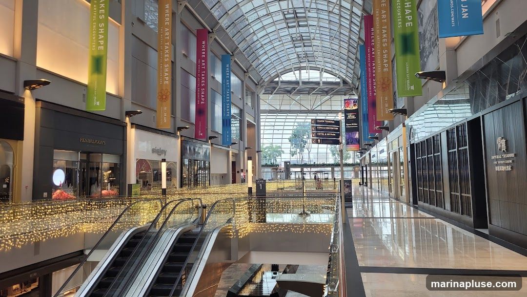 An interior view of a modern shopping mall with escalators, decorative lights, and large banners hanging from the ceiling. Several storefronts are visible, including Penhaligon's and Imperial Treasure Fine Chinese Cuisine. Directional signs indicate various destinations like Coach Terminal, Expo & Convention, and Hotel & Skypark.