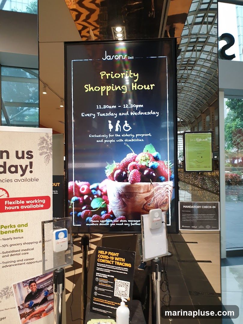 A digital screen displays an advertisement for Jasons Deli's Priority Shopping Hour, which is held every Tuesday and Wednesday from 11:30 am to 12:30 pm, exclusively for the elderly, pregnant, and people with disabilities. The screen also shows an image of fresh berries. To the left, a poster advertises flexible working hours and perks. Below the digital screen, there is a sign for COVID-19 contact tracing and a hand sanitizer dispenser.